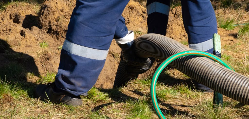 Man worker holding pipe, providing sewer cleaning service outdoor. Sewage pumping machine is unclogging blocked manhole